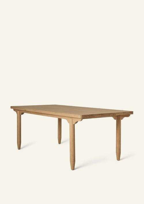 Three-quarter view of the dining table in White Oak. Crafted from solid oak, the table features softly rounded corners, sculptural pencil legs, and scalloped apron detailing that lends a collected, timeless character.
