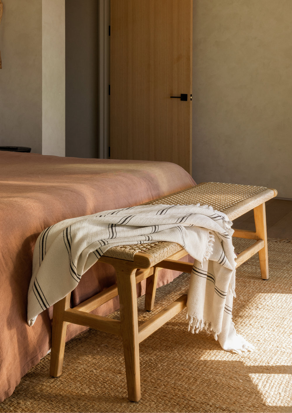 Teak bench positioned at the foot of a bed and styled with a draped throw blanket. Warm natural materials and soft sunlight create an inviting bedroom setting that highlights the bench’s versatility.