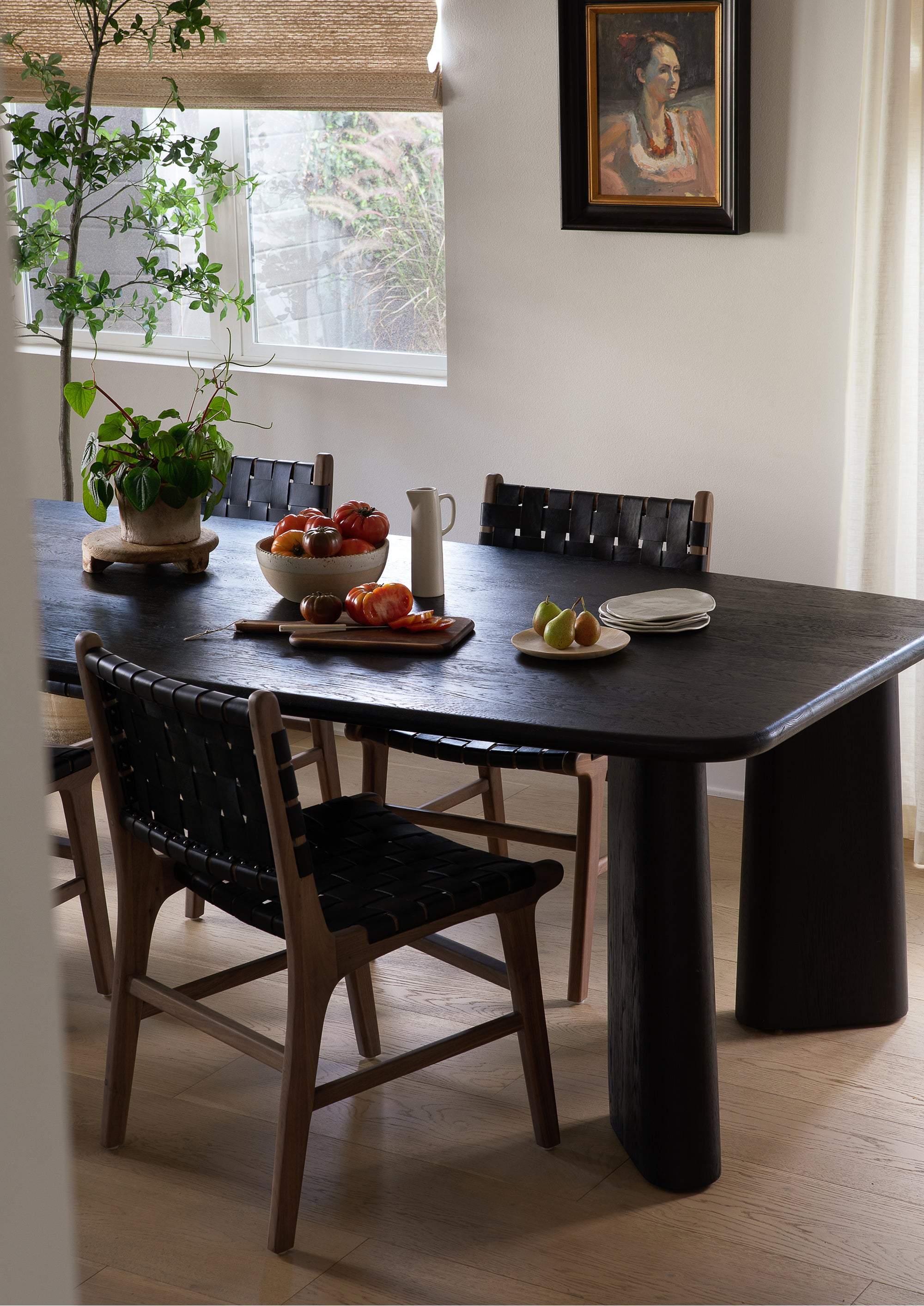 dining space featuring multiple Bali Dining Chairs with woven black leather seats and backs gathered around a sculptural dark wood dining table. Natural light and layered textures create a sophisticated, welcoming atmosphere.