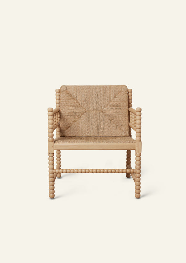 Front view of a natural oak lounge chair with a woven rush seat and geometric woven backrest. Turned spindle details frame the arms and legs, creating a sculptural silhouette with artisanal character. Photographed against a warm ivory backdrop.