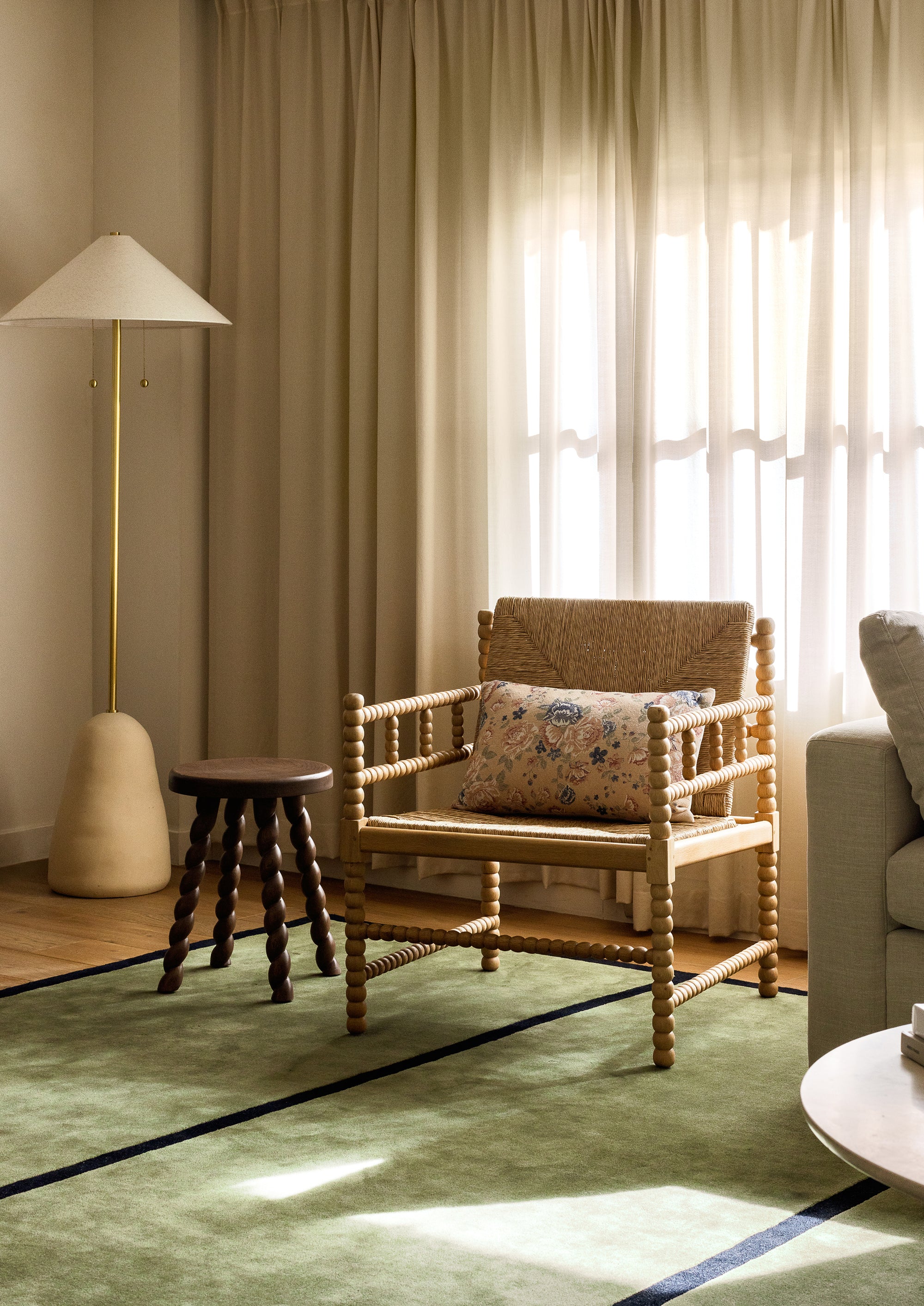 Natural oak lounge chair with woven rush seat and backrest styled in a sunlit living room. A floral pillow, green area rug, sculptural floor lamp, and dark wood side table create a warm, collected seating vignette.