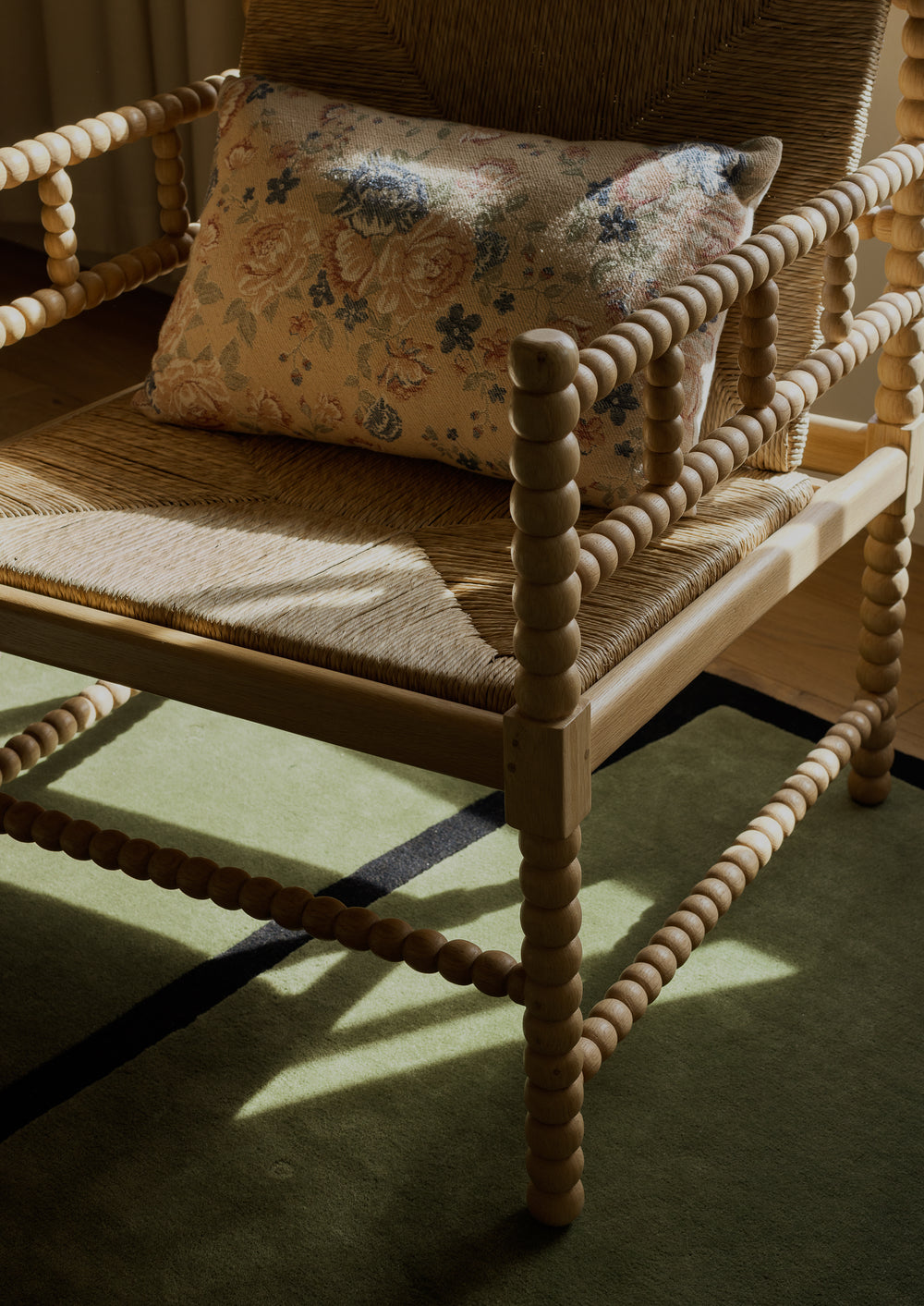Close-up of a natural oak lounge chair with woven rush details and turned spindle arms. A floral accent pillow rests against the backrest as sunlight casts soft shadows across the chair and surrounding rug.