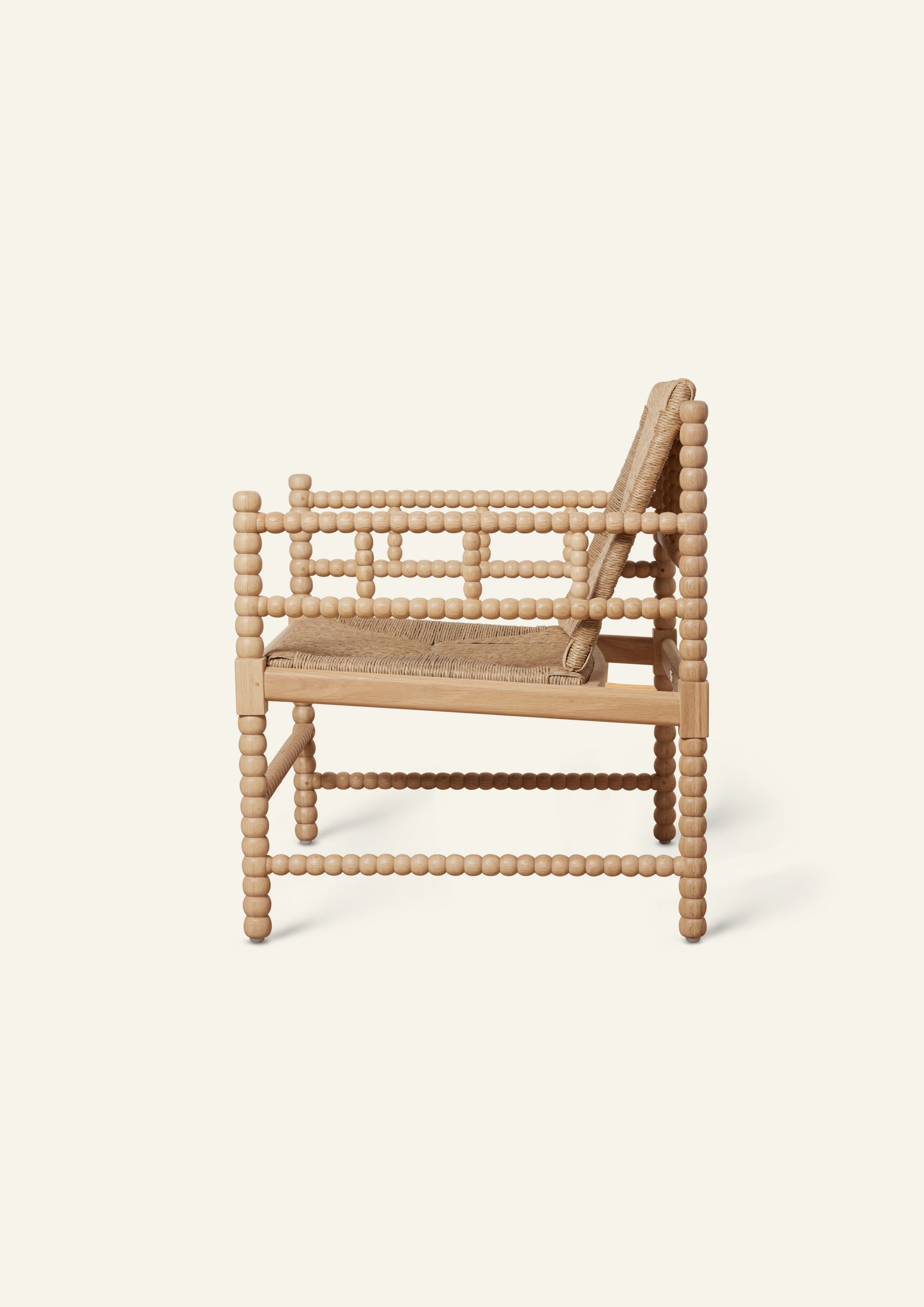 Side profile of a natural oak lounge chair featuring a handwoven rush seat and backrest. Decorative turned spindle detailing runs along the arms, legs, and stretcher supports, highlighting the chair’s handcrafted construction.