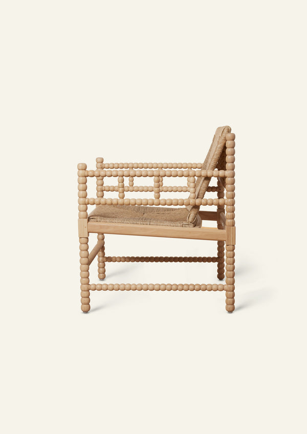 Side profile of a natural oak lounge chair featuring a handwoven rush seat and backrest. Decorative turned spindle detailing runs along the arms, legs, and stretcher supports, highlighting the chair’s handcrafted construction.
