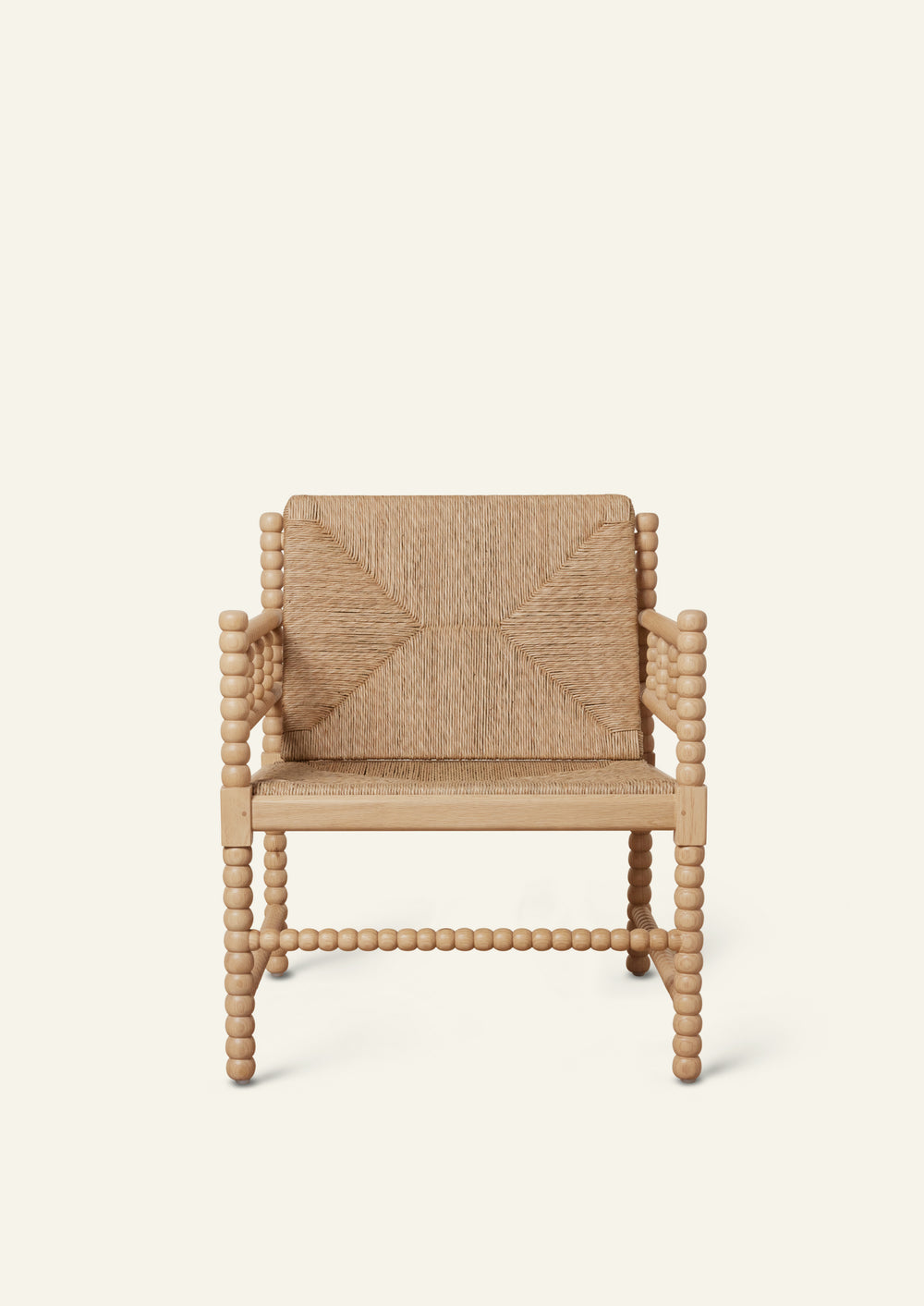 Front view of a natural oak lounge chair with a woven rush seat and geometric woven backrest. Turned spindle details frame the arms and legs, creating a sculptural silhouette with artisanal character. Photographed against a warm ivory backdrop.
