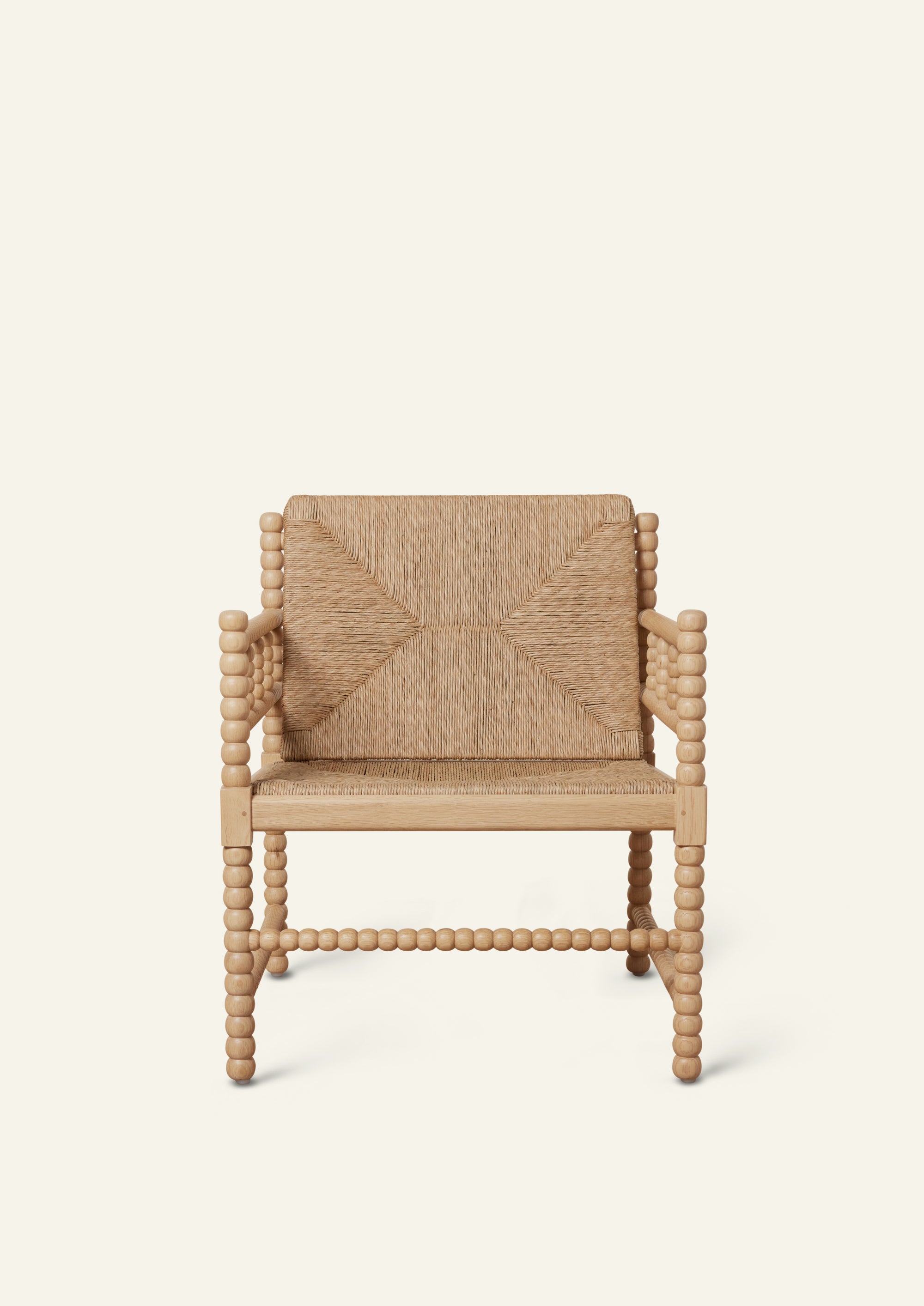 Front view of a natural oak lounge chair with a woven rush seat and geometric woven backrest. Turned spindle details frame the arms and legs, creating a sculptural silhouette with artisanal character. Photographed against a warm ivory backdrop.