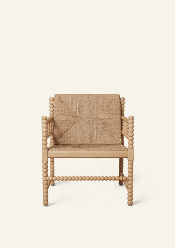 Front view of a natural oak lounge chair with a woven rush seat and geometric woven backrest. Turned spindle details frame the arms and legs, creating a sculptural silhouette with artisanal character. Photographed against a warm ivory backdrop.