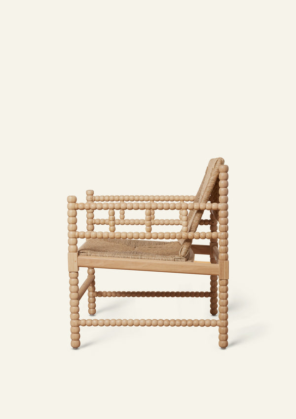 Side profile of a natural oak lounge chair featuring a handwoven rush seat and backrest. Decorative turned spindle detailing runs along the arms, legs, and stretcher supports, highlighting the chair’s handcrafted construction.
