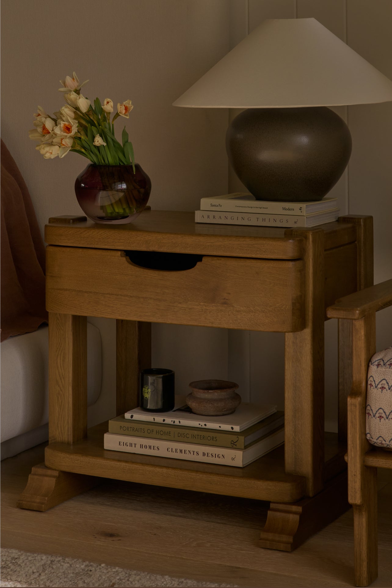 Wooden nightstand with books, a lamp, and a vase of flowers on top.