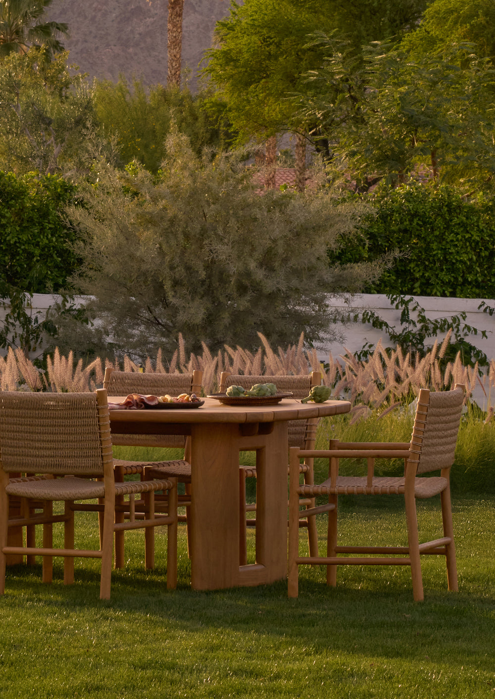 A collection of woven teak armchairs surrounding an outdoor dining table on a landscaped lawn. The durable materials and handcrafted details blend seamlessly with the natural environment.
