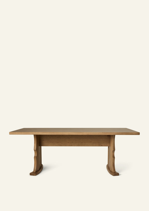 Side profile of the trestle dining table highlighting the sculptural pedestal base, center stretcher, and substantial tabletop. The architectural design balances strength and elegance while maximizing seating flexibility.