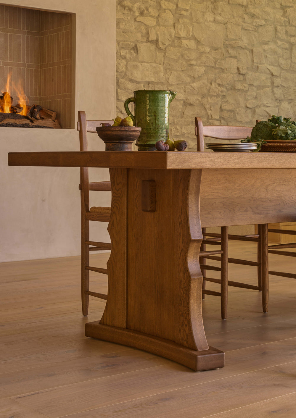 Close-up of the trestle dining table styled in a dining room with woven-seat chairs, ceramic serveware, and seasonal produce. The warm Honey Oak finish and architectural base create a collected, welcoming atmosphere.