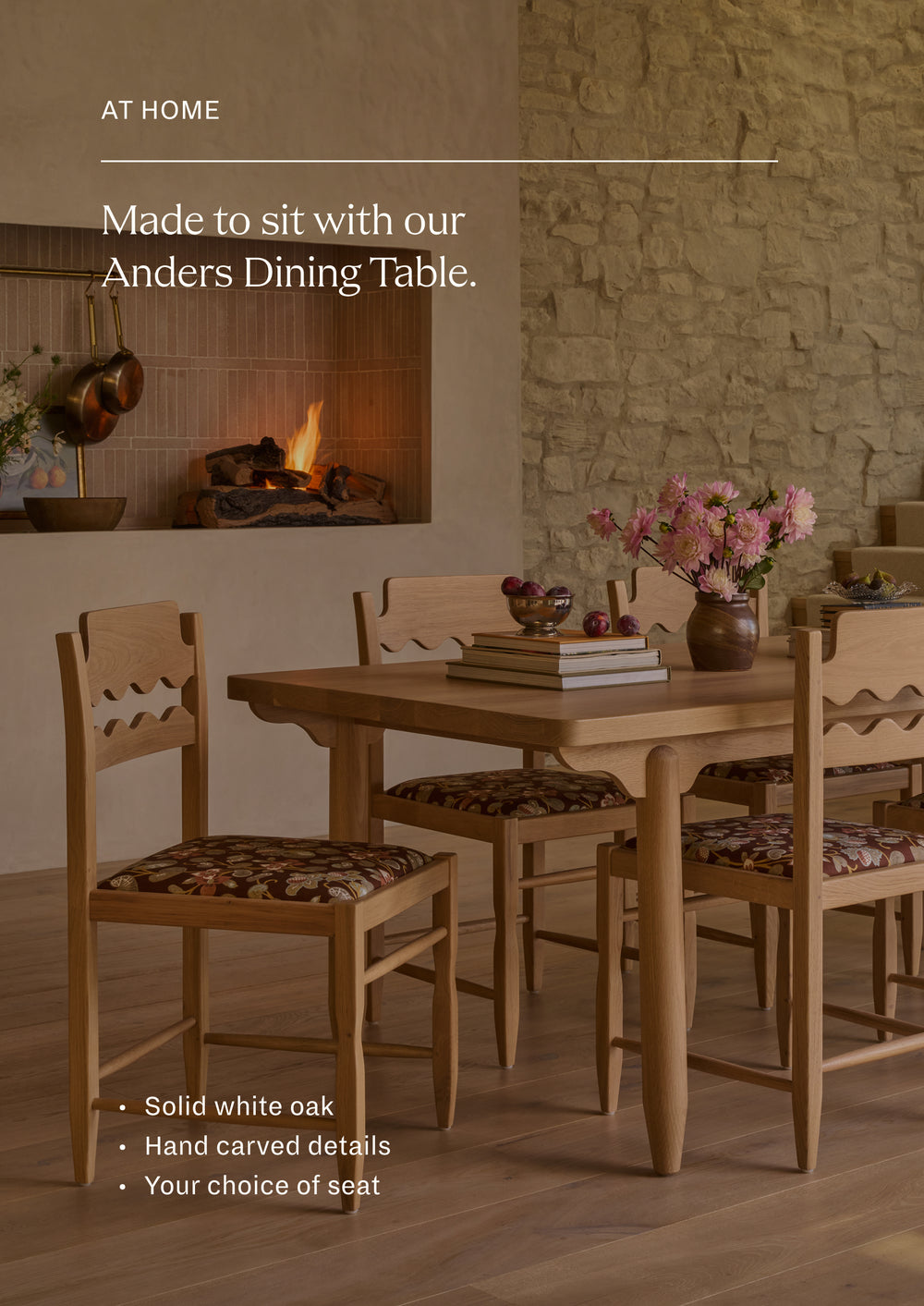 Freda Razor Dining Chairs gathered around the white oak Anders Dining Table in a warm, stone-walled dining room. The chairs feature hand-carved scalloped back details, upholstered floral seats, and a light oak finish styled with books, flowers, and tableware.