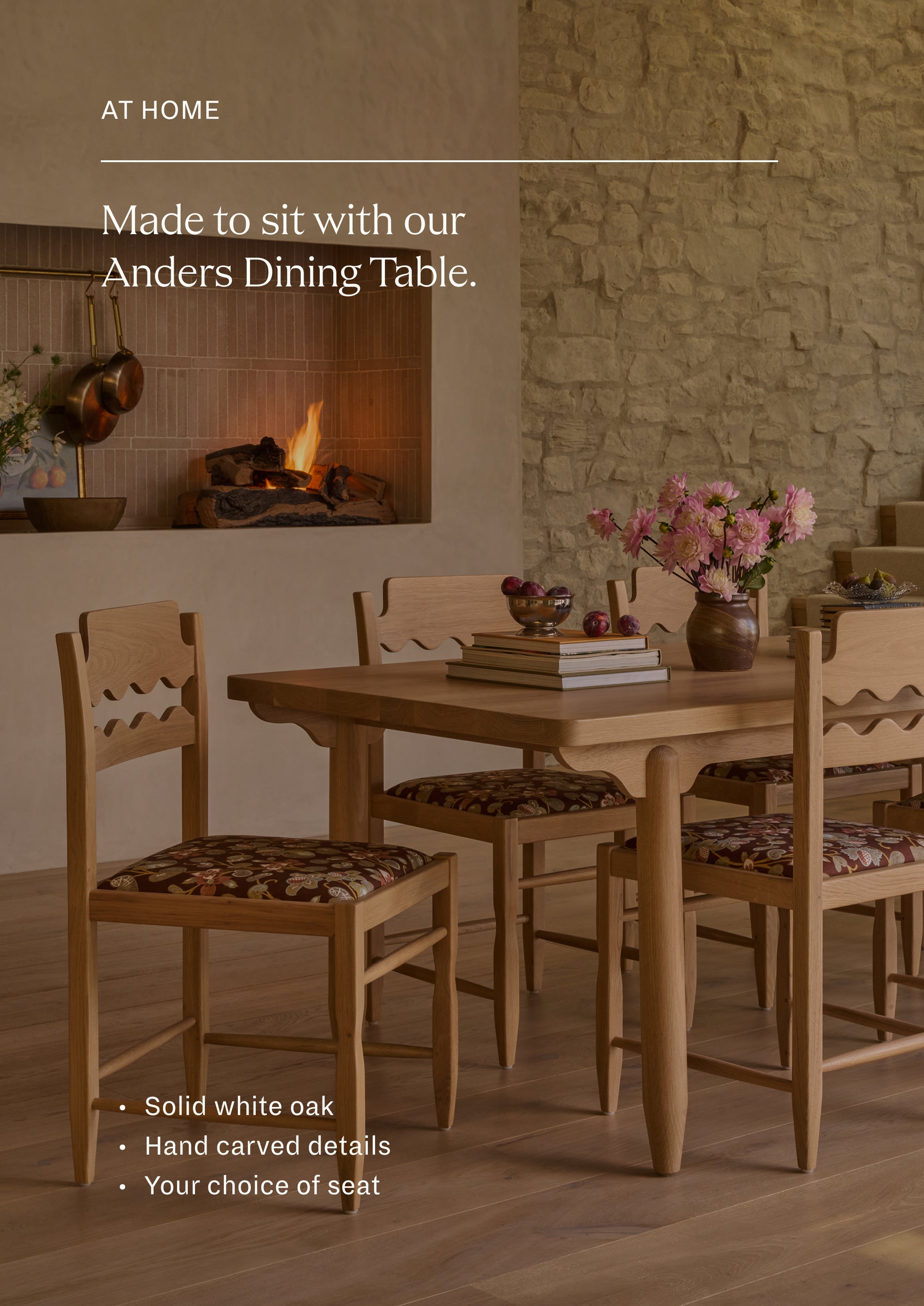 Freda Razor Dining Chairs gathered around the white oak Anders Dining Table in a warm, stone-walled dining room. The chairs feature hand-carved scalloped back details, upholstered floral seats, and a light oak finish styled with books, flowers, and tableware.