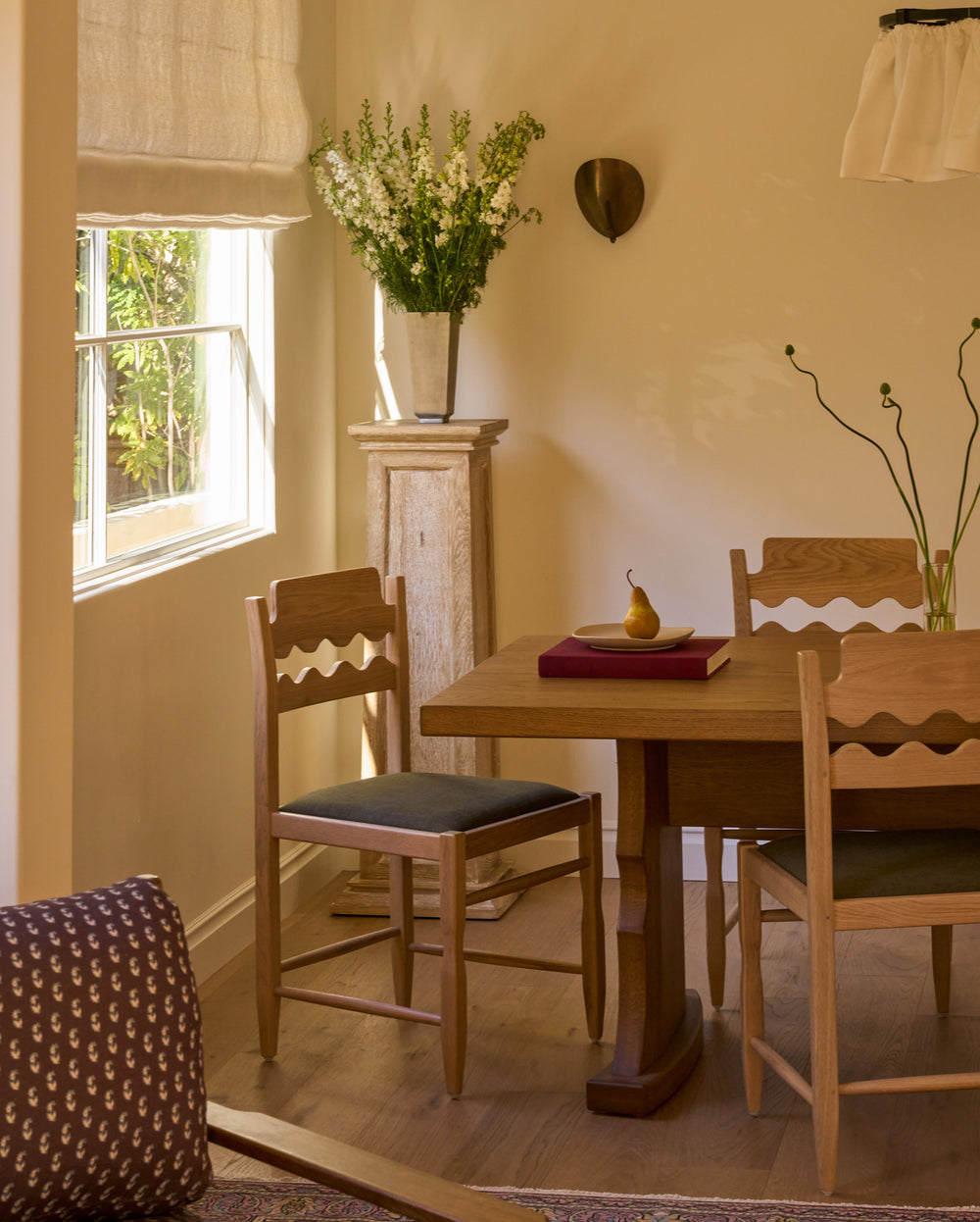 Freda Razor Dining Chairs with olive upholstered seats surrounding the Etta honey oak dining table in a softly lit dining room. The hand-carved scalloped backrests and natural oak finish create a timeless, European-inspired look.