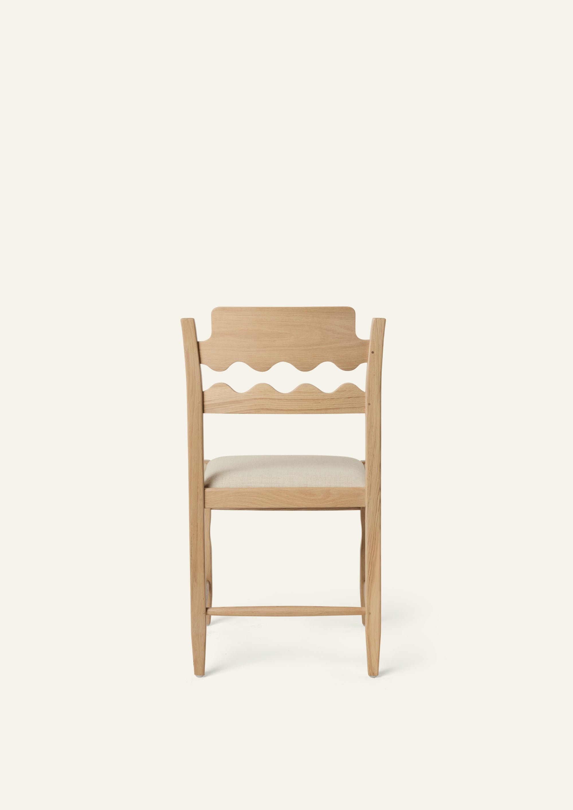 Back view of the Freda Razor Dining Chair in natural oak with an upholstered performance sand linen seat, shown from an angled front view on a neutral background. The hand-carved scalloped backrest adds a playful yet timeless detail.