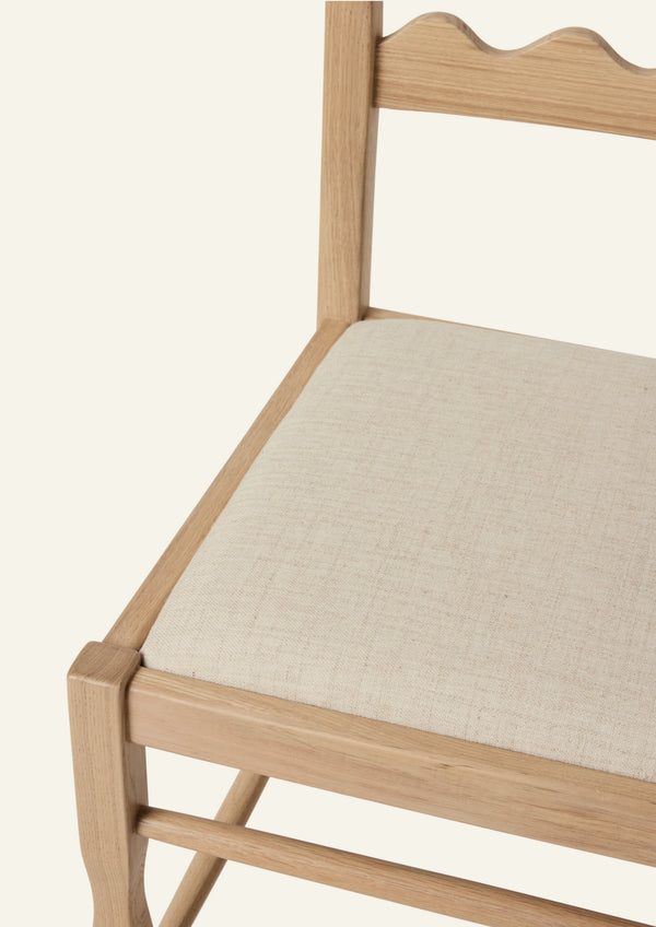 Detailed view of the Freda Razor Dining Chair in natural oak with an upholstered performance sand linen seat, shown from an angled front view on a neutral background. The hand-carved scalloped backrest adds a playful yet timeless detail.