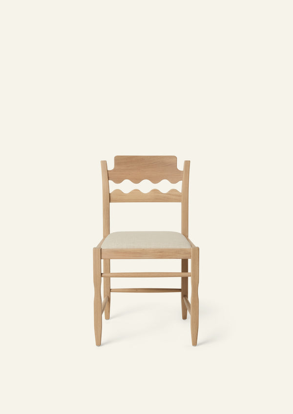 Front view of the Freda Razor Dining Chair in natural oak with an upholstered performance sand linen seat, shown from an angled front view on a neutral background. The hand-carved scalloped backrest adds a playful yet timeless detail.