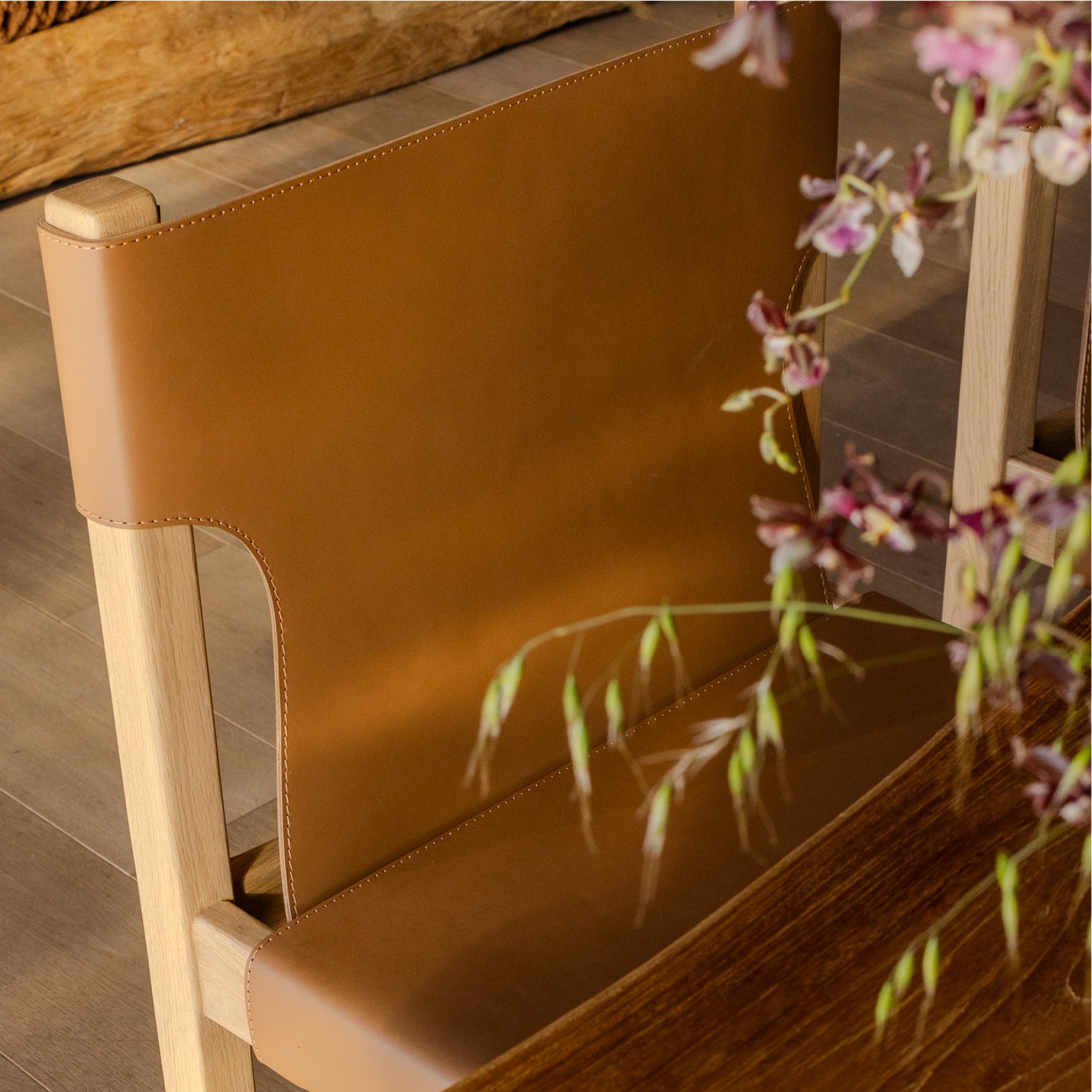 Brown leather chair with white oak legs on a wooden floor, accompanied by a plant in the foreground.