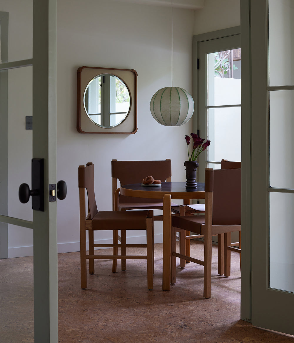 Small, modern dining nook with wooden table and Henrik leather dining chairs, round mirror, and pendant light.