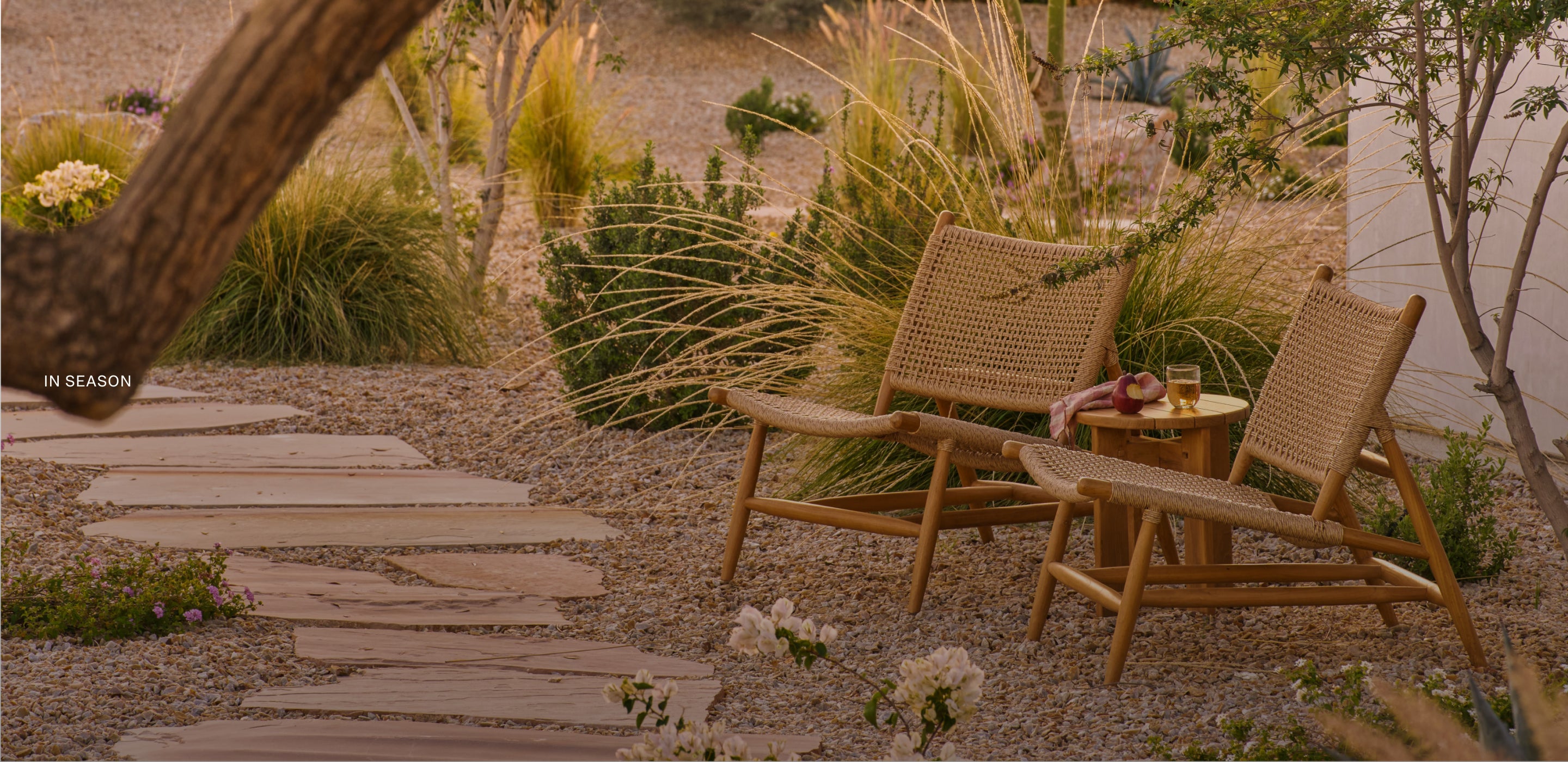 Two synthetic lounge (Bali Lounge) chairs and a small table (Otto Table) in a garden setting with plants and flowers.