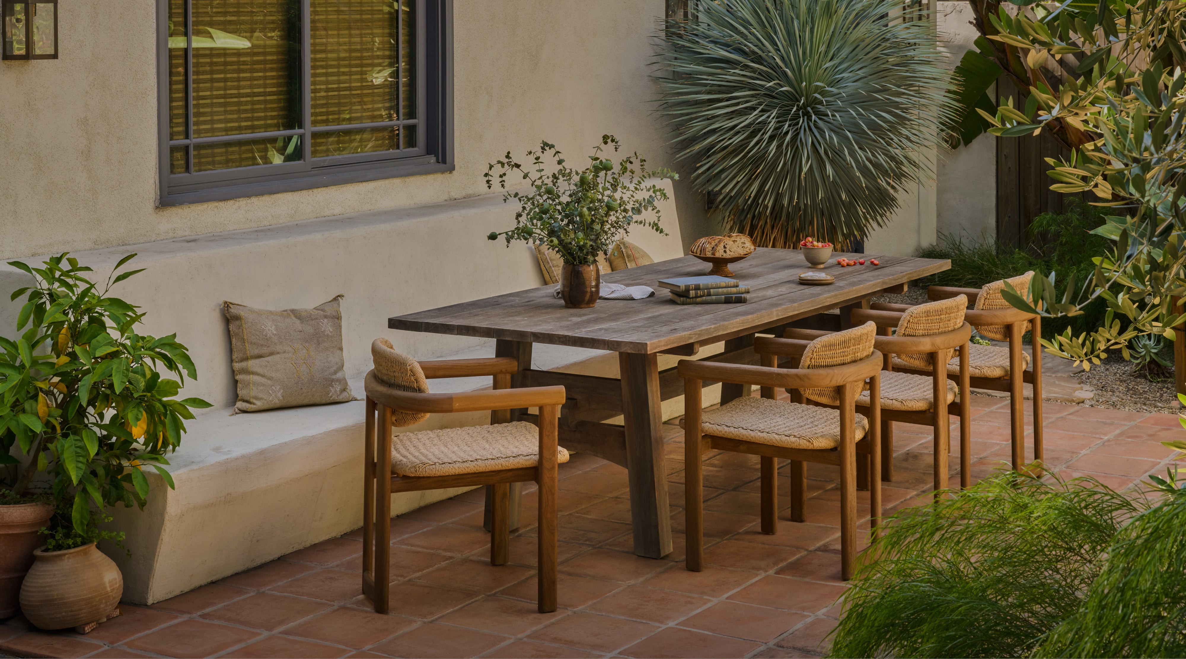 Outdoor patio setting with wooden teak table and Vera teak dining chairs surrounded by plants on a terra cotta patio. 