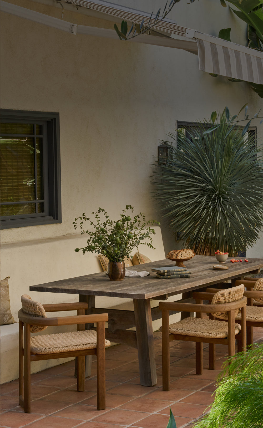 Outdoor patio setting with wooden teak table and Vera teak dining chairs surrounded by plants on a terra cotta patio. 