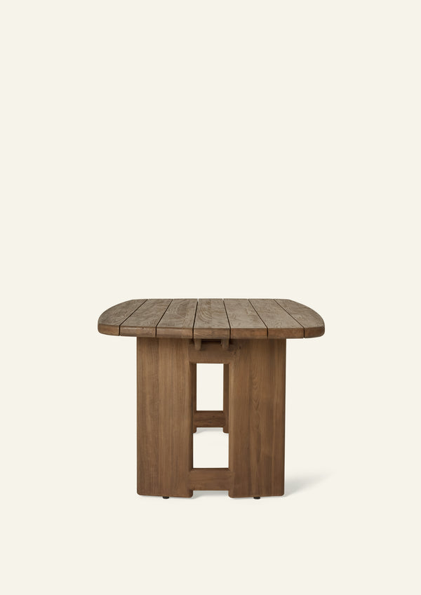 Side profile of the reclaimed teak dining table highlighting the slim tabletop, rounded edges, and architectural pedestal supports. The slatted construction allows rainwater to drain through while maintaining a refined, sculptural appearance.
