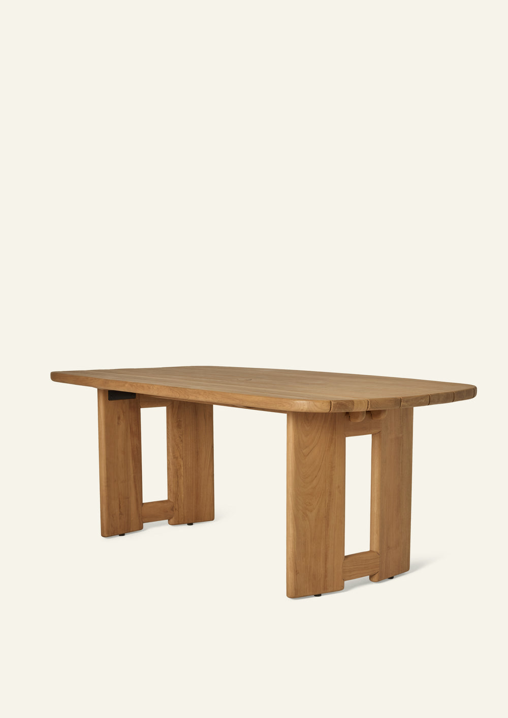 Three-quarter view of an outdoor dining table crafted from solid teak. The slatted tabletop features softly rounded corners and a substantial architectural base that creates a grounded, sculptural presence while allowing rainwater to drain through.