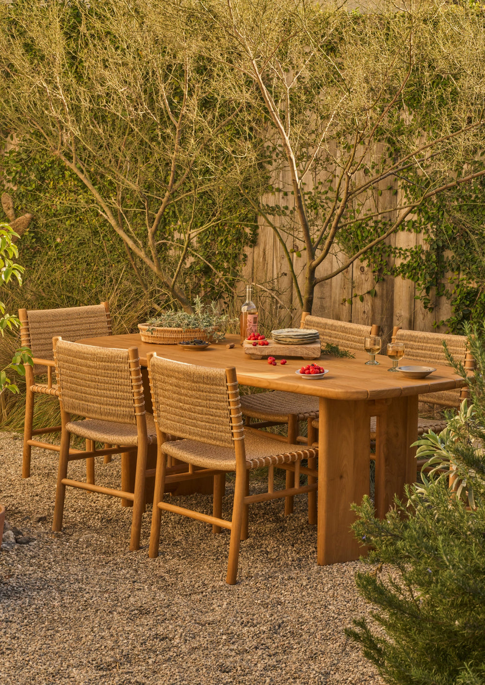 The teak dining table styled in a garden setting with matching woven chairs. Surrounded by natural landscaping, the table serves as a warm and welcoming gathering place for outdoor dining and conversation.