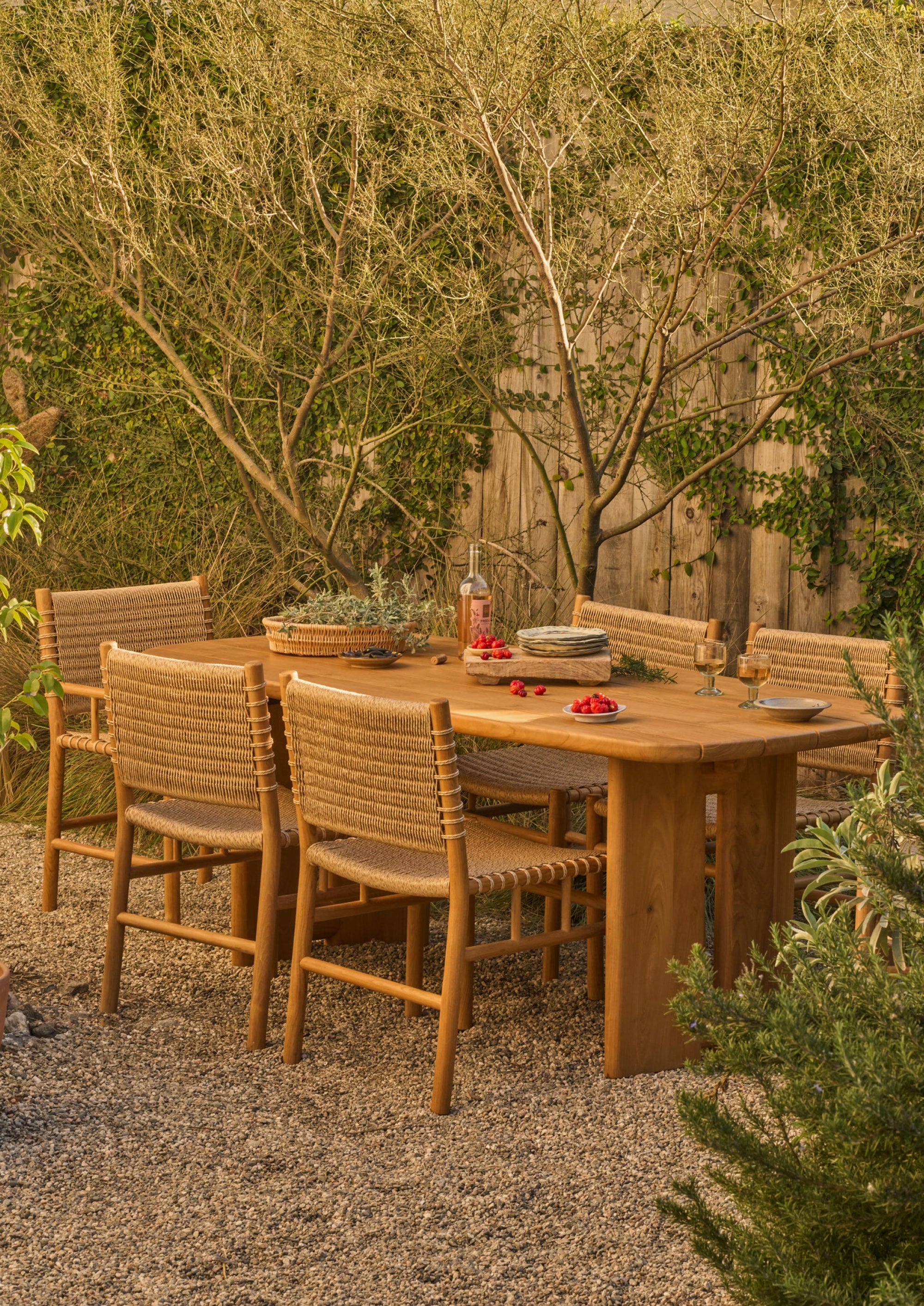The teak dining table styled in a garden setting with matching woven chairs. Surrounded by natural landscaping, the table serves as a warm and welcoming gathering place for outdoor dining and conversation.