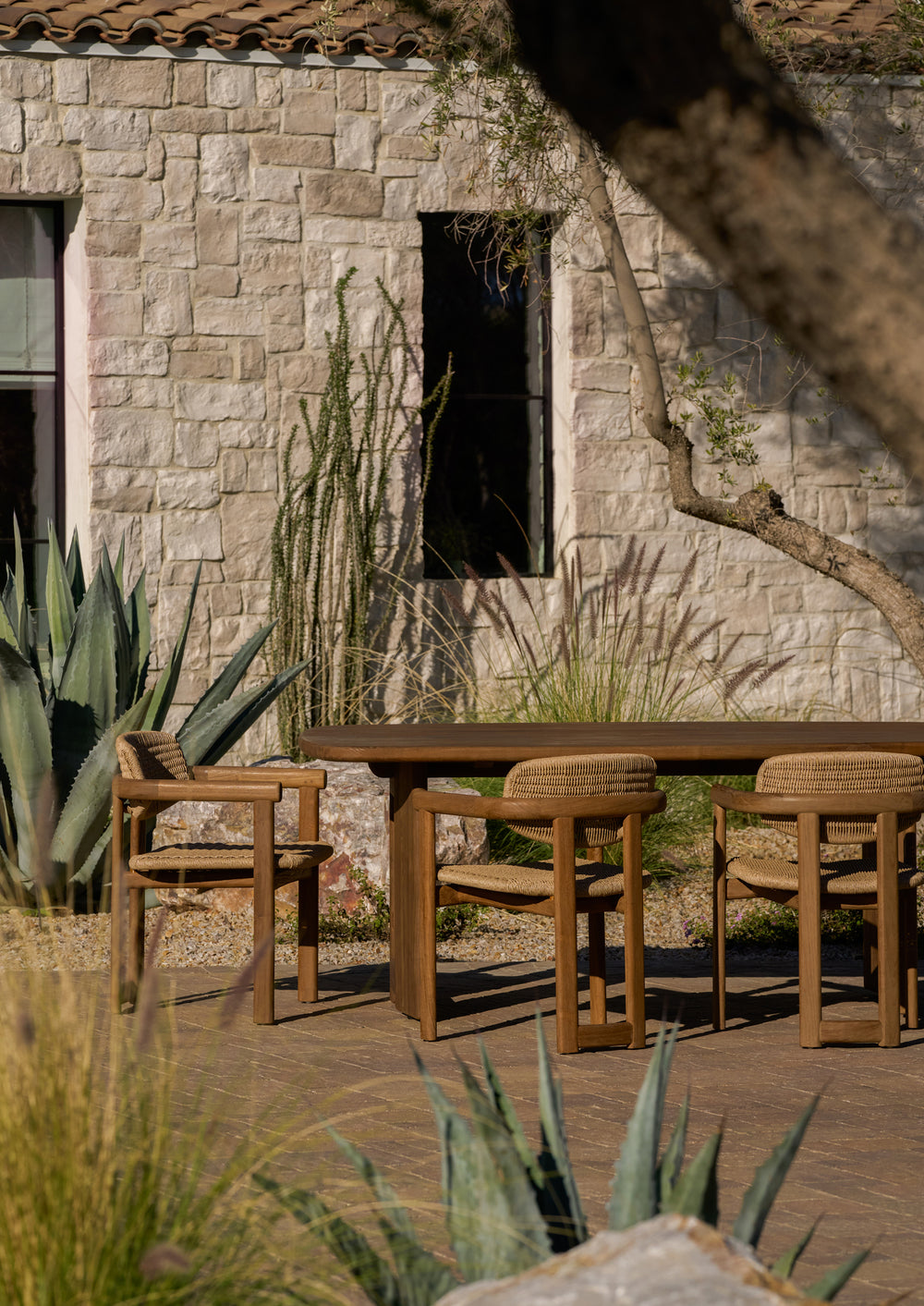 The reclaimed teak dining table styled on a stone patio with coordinating outdoor dining chairs. Surrounded by desert-inspired landscaping, the table anchors an inviting outdoor dining space designed for everyday living.