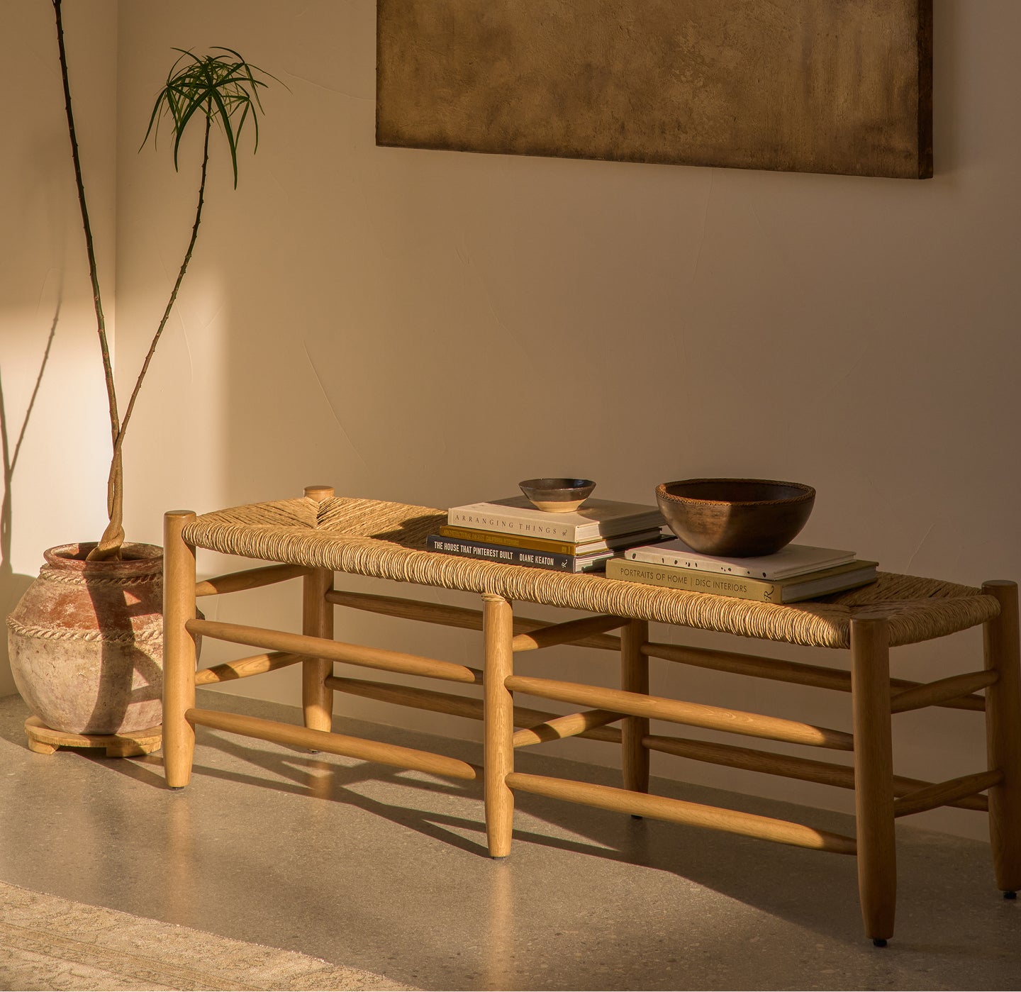 Mina honey oak bench with woven  rush seat in a room with a plant and painting on the wall.