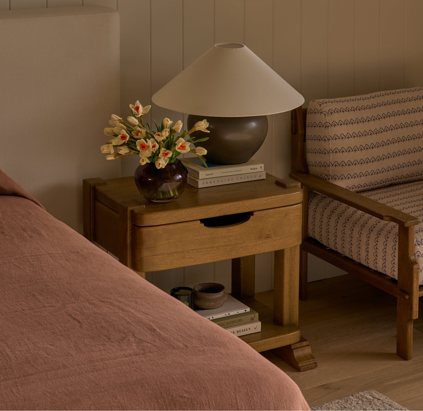 Mina honey oak nightstand with lamp, books, and flowers next to a bed in a bedroom setting.