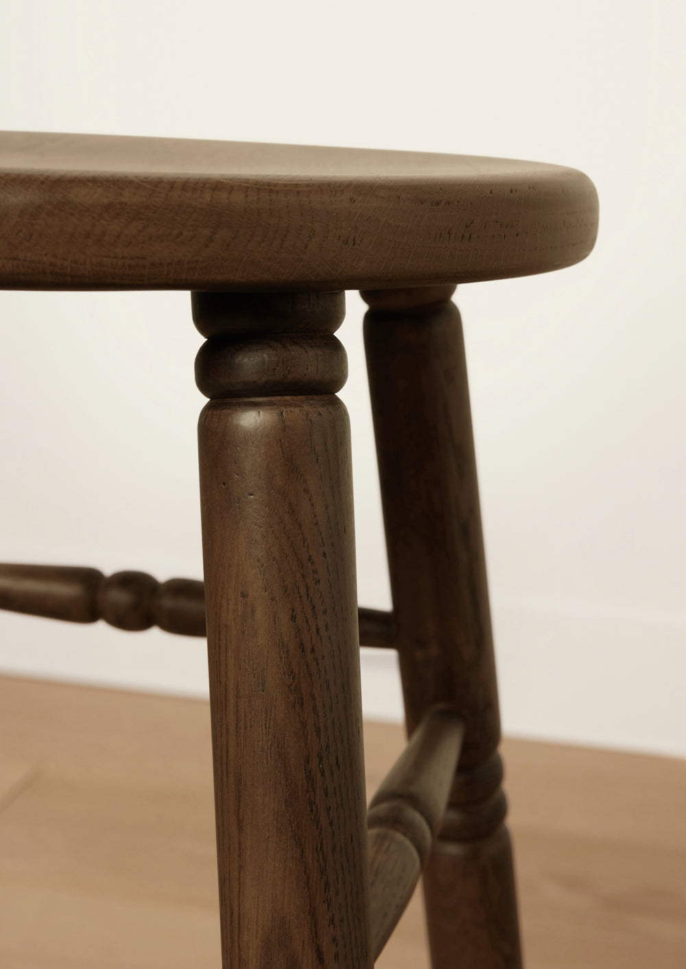 Side profile of the backless counter stool featuring a round oak seat, tapered turned legs, and connecting stretchers. The traditional form is elevated by its refined proportions and warm brown finish.