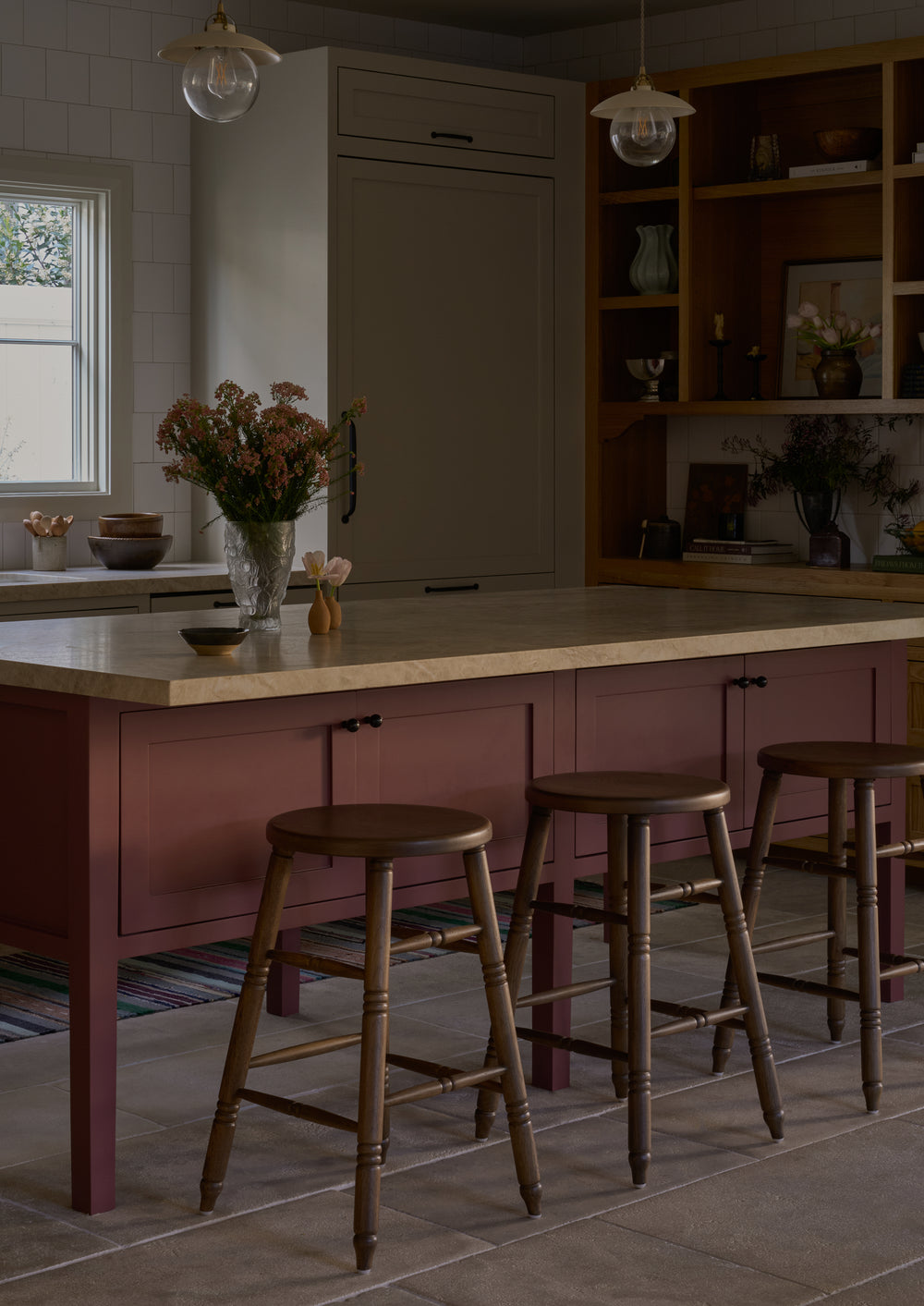 Three brown oak counter stools with hand-turned details are arranged around a mauve kitchen island topped with a light stone countertop. The stools bring warmth and traditional character to the space, complemented by open wood shelving, soft lighting, and fresh floral arrangements.