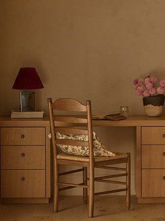 Wooden desk with Lea Dining chair in brown oak, lamp, and flowers against a plain wall.