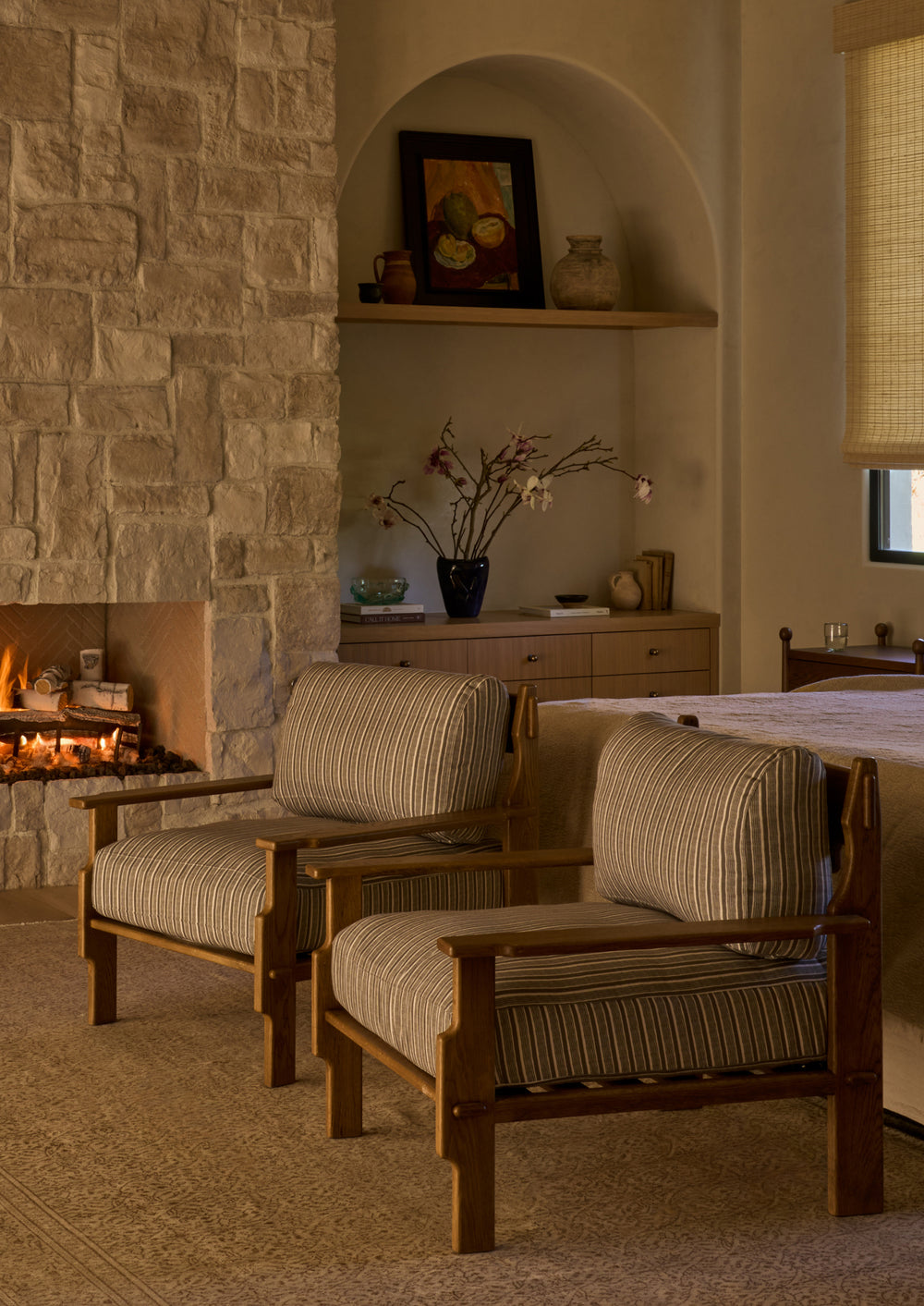 Two lounge chairs upholstered in green stripe fabric are arranged beside a stone fireplace in a warm, inviting bedroom. The striped upholstery and natural oak frames create a relaxed, timeless atmosphere.