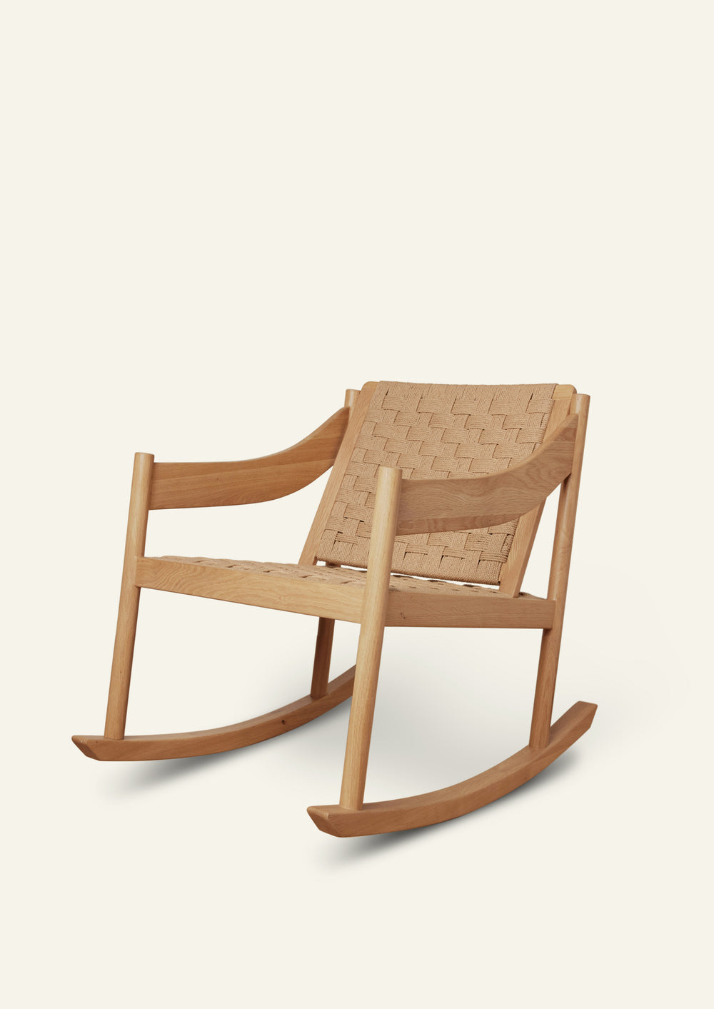 Natural oak rocking chair with a woven paper cord seat and backrest, featuring gently curved armrests and smooth rocker runners. Shown at a three-quarter angle against a soft ivory background.