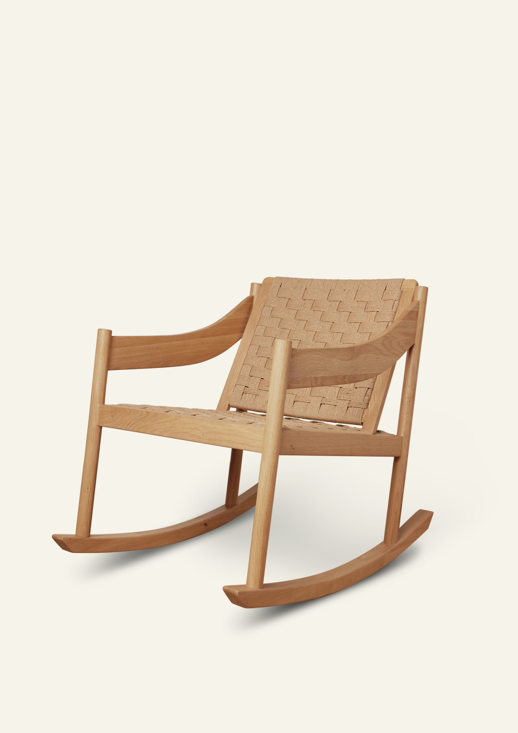 Natural oak rocking chair with a woven paper cord seat and backrest, featuring gently curved armrests and smooth rocker runners. Shown at a three-quarter angle against a soft ivory background.