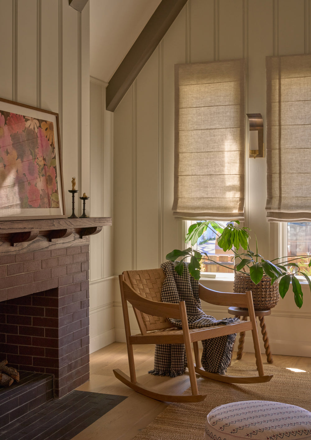 Natural oak rocking chair with a woven paper cord seat and backrest styled in a cozy living room. A checkered throw blanket drapes over the chair beside a side table, houseplant, and brick fireplace, creating a warm and inviting reading corner.