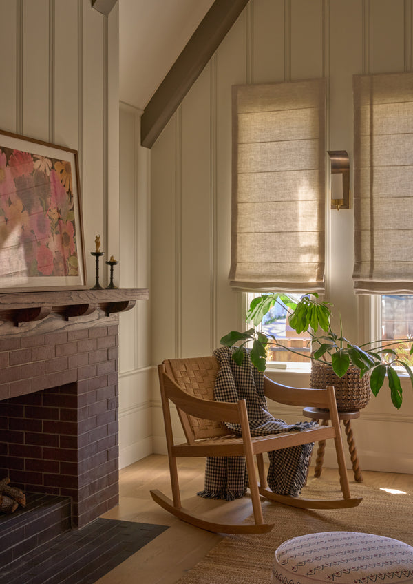 Natural oak rocking chair with a woven paper cord seat and backrest styled in a cozy living room. A checkered throw blanket drapes over the chair beside a side table, houseplant, and brick fireplace, creating a warm and inviting reading corner.