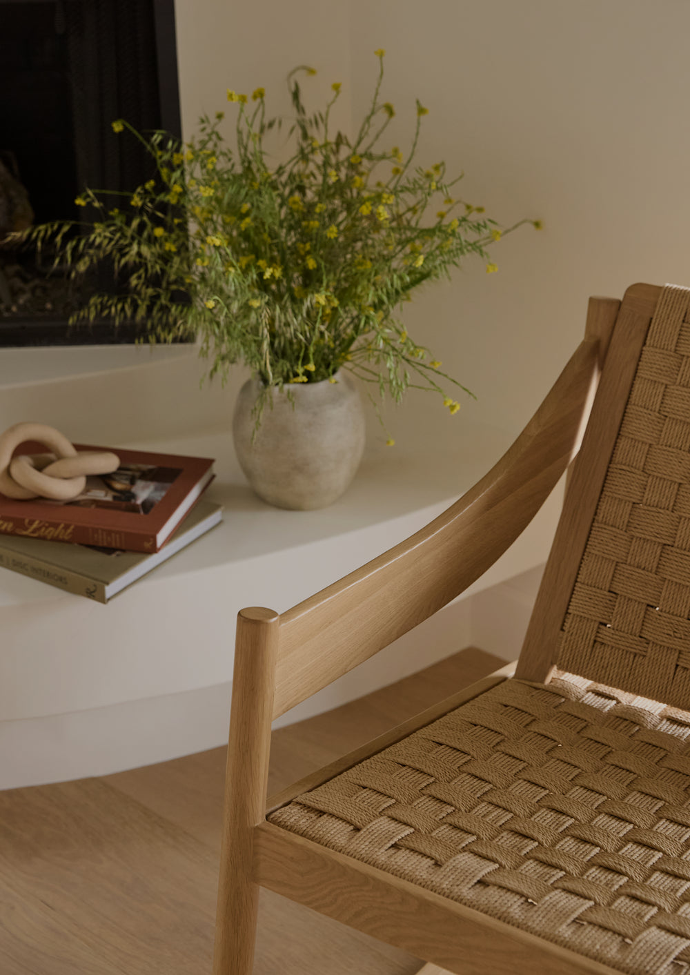 Close-up of a natural oak rocking chair with woven paper cord detailing. Warm sunlight highlights the sculpted wood armrest and textured weave, while a vase of fresh flowers and stacked books sit nearby.