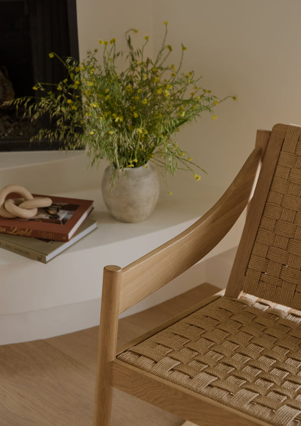 Close-up of a natural oak rocking chair with woven paper cord detailing. Warm sunlight highlights the sculpted wood armrest and textured weave, while a vase of fresh flowers and stacked books sit nearby.