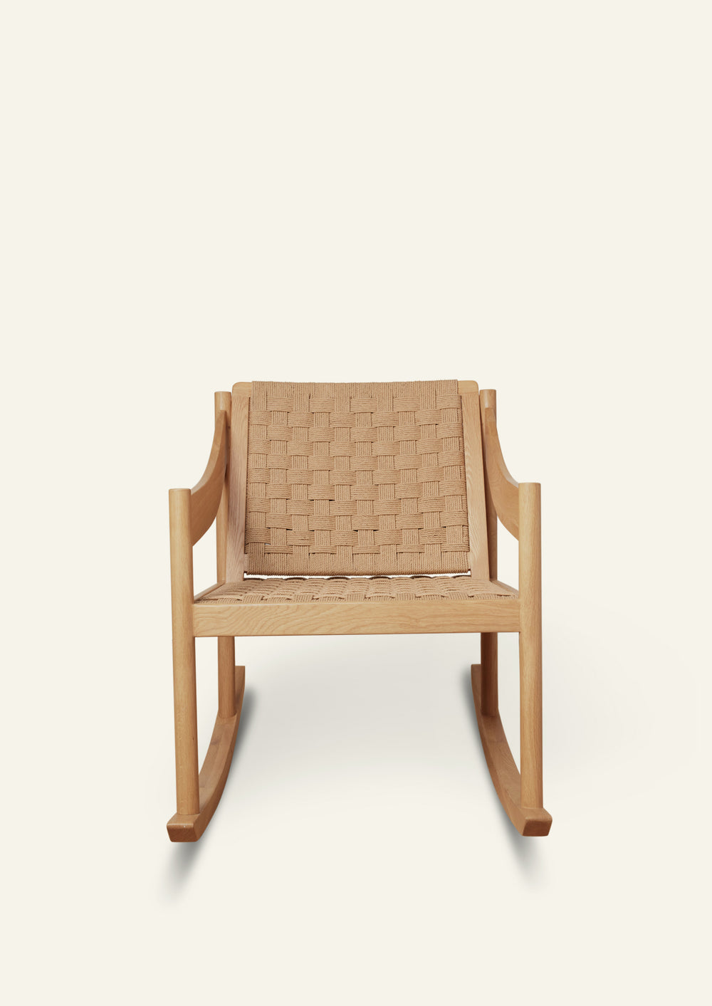 Front view of a natural oak rocking chair with a handwoven paper cord seat and backrest. The chair features clean lines, curved armrests, and a relaxed silhouette designed for comfort. Photographed against a warm ivory backdrop.