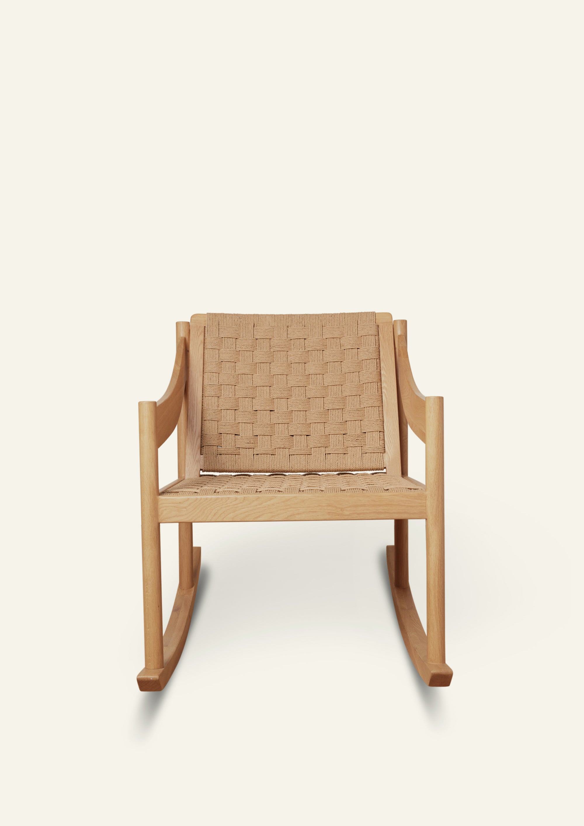 Front view of a natural oak rocking chair with a handwoven paper cord seat and backrest. The chair features clean lines, curved armrests, and a relaxed silhouette designed for comfort. Photographed against a warm ivory backdrop.