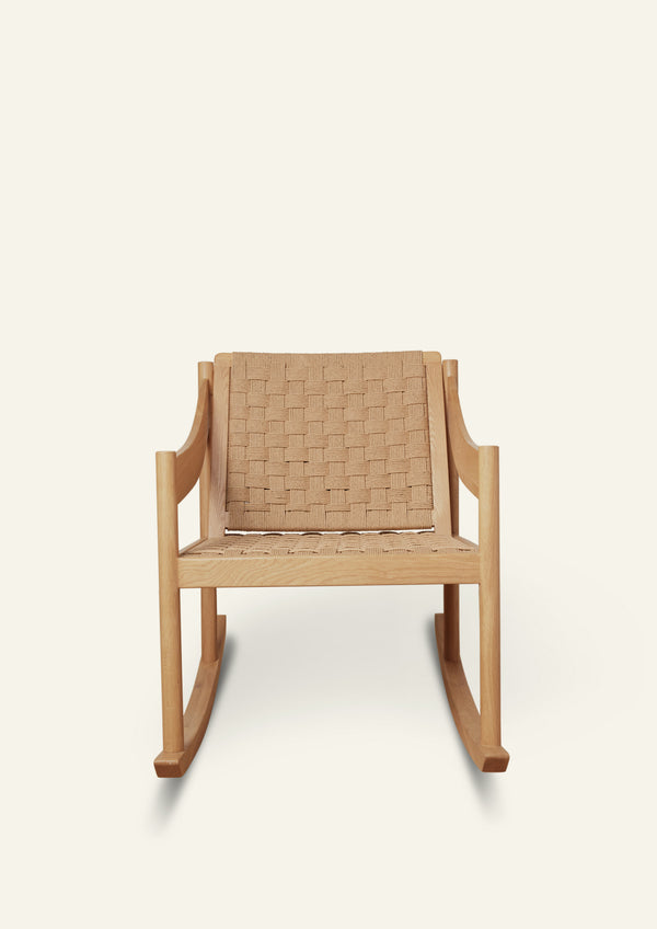 Front view of a natural oak rocking chair with a handwoven paper cord seat and backrest. The chair features clean lines, curved armrests, and a relaxed silhouette designed for comfort. Photographed against a warm ivory backdrop.