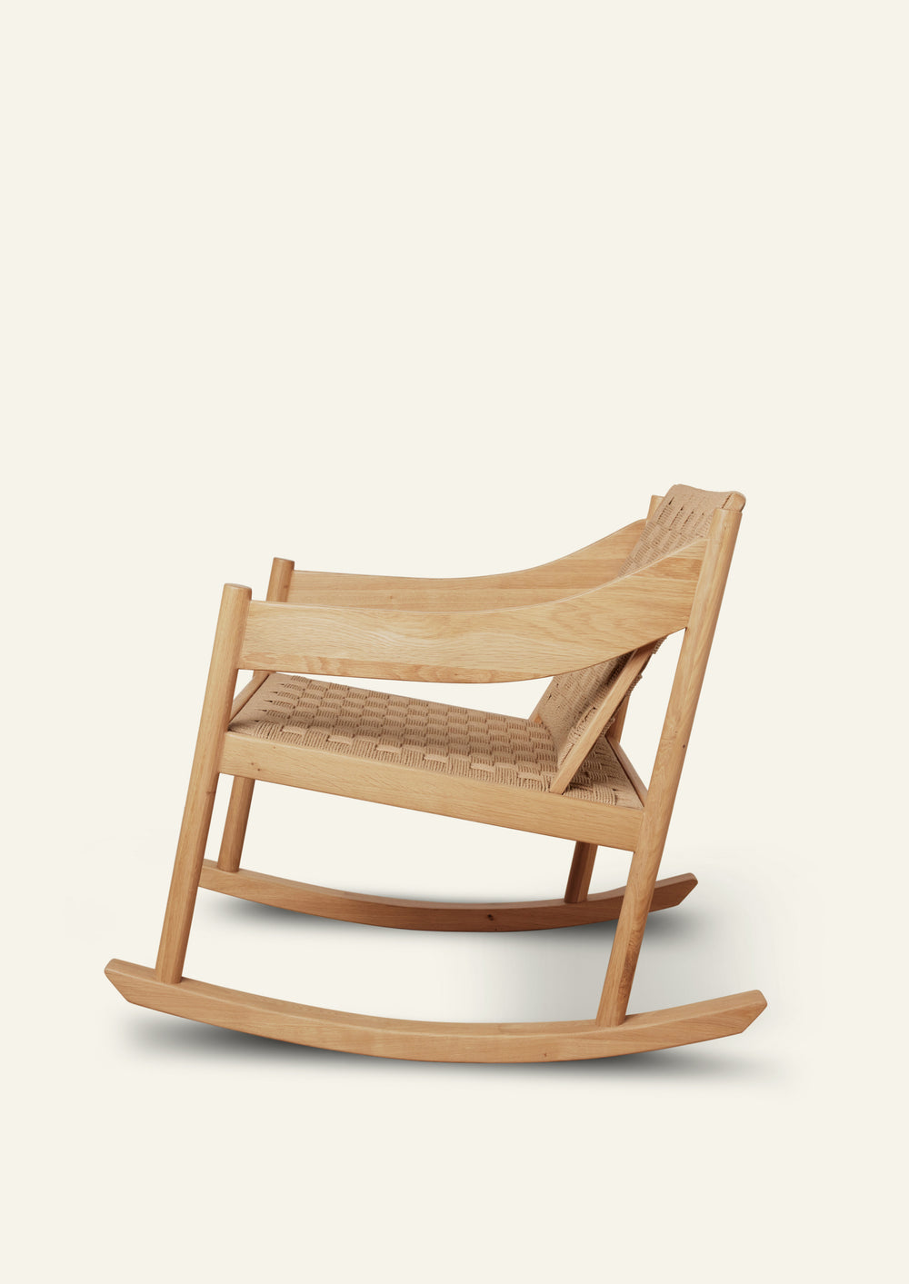 Side profile of a natural oak rocking chair featuring a woven paper cord seat and backrest. Curved rocker runners and sculpted armrests create a balanced silhouette with Scandinavian-inspired simplicity.