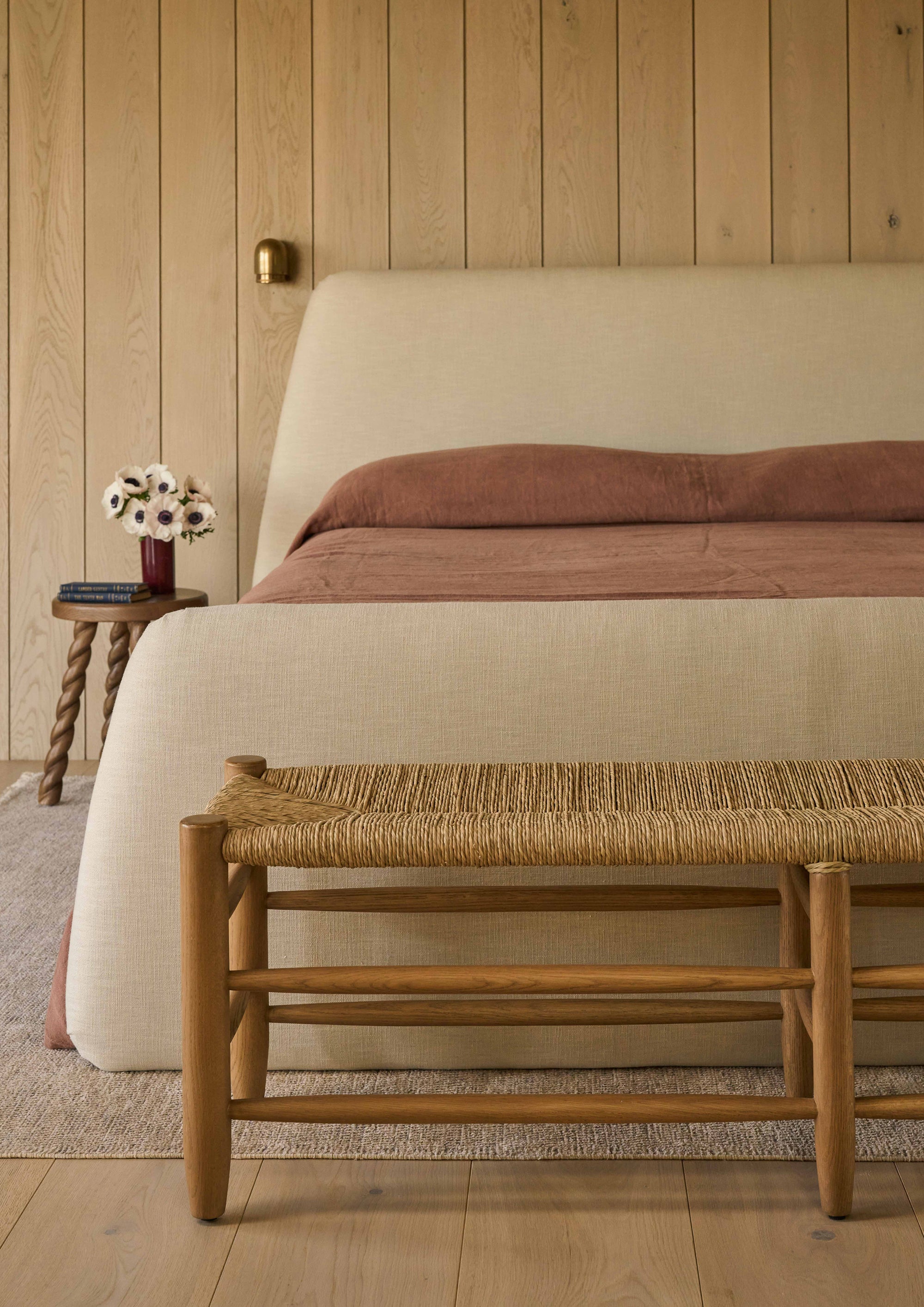 Honey oak bench placed at the foot of an upholstered bed, styled within a warm and inviting bedroom. The woven rush seat and natural oak frame add texture, warmth, and casual elegance to the space.