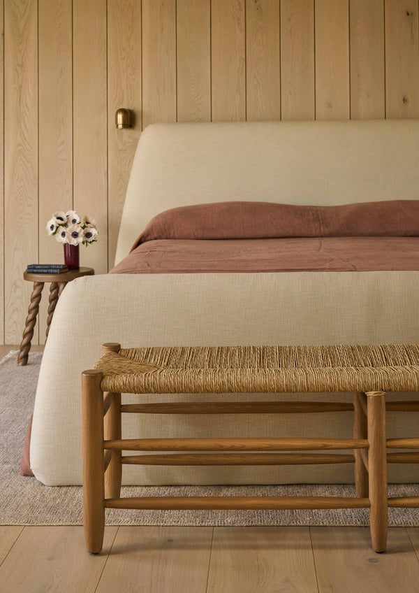 Honey oak bench placed at the foot of an upholstered bed, styled within a warm and inviting bedroom. The woven rush seat and natural oak frame add texture, warmth, and casual elegance to the space.